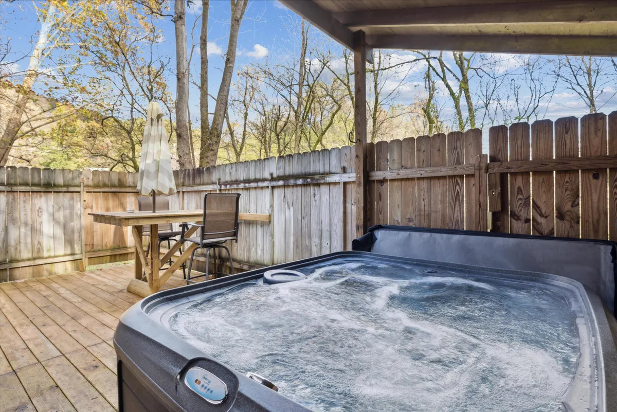 A tranquil outdoor deck featuring a bubbling hot tub surrounded by trees. In the scene, there is also a dining table with chairs, making it an inviting space for both relaxation and outdoor meals. The area feels private and nestled within nature, perfect for peaceful evenings or mornings.