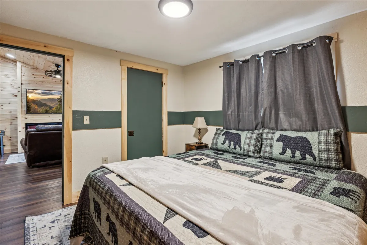 A rustic bedroom with a bear-themed bedspread, a window draped with curtains letting in natural light, and a bedside lamp. The space feels warm, comfortable, and perfect for reading or reflecting in solitude.