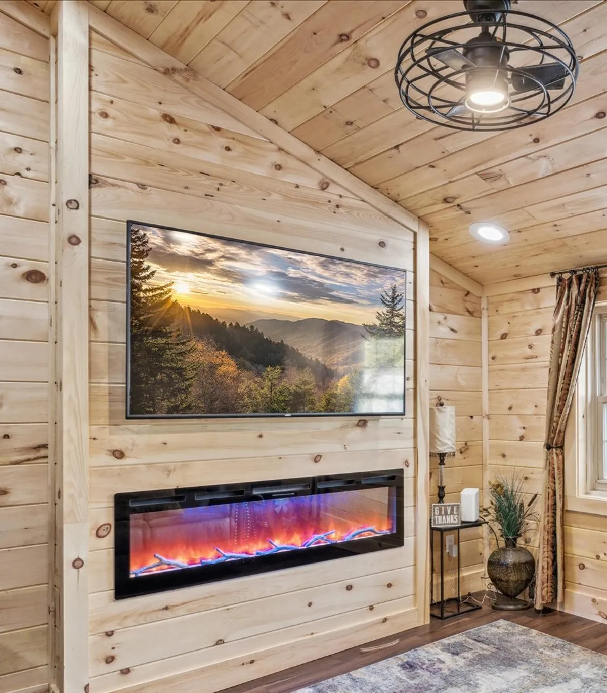 A cozy living room with wooden paneling, a glowing electric fireplace beneath framed landscape artwork, a decorative lamp, and a potted plant. Plush seating invites relaxation and the ambiance is warm and rustic, evoking a mountain retreat.