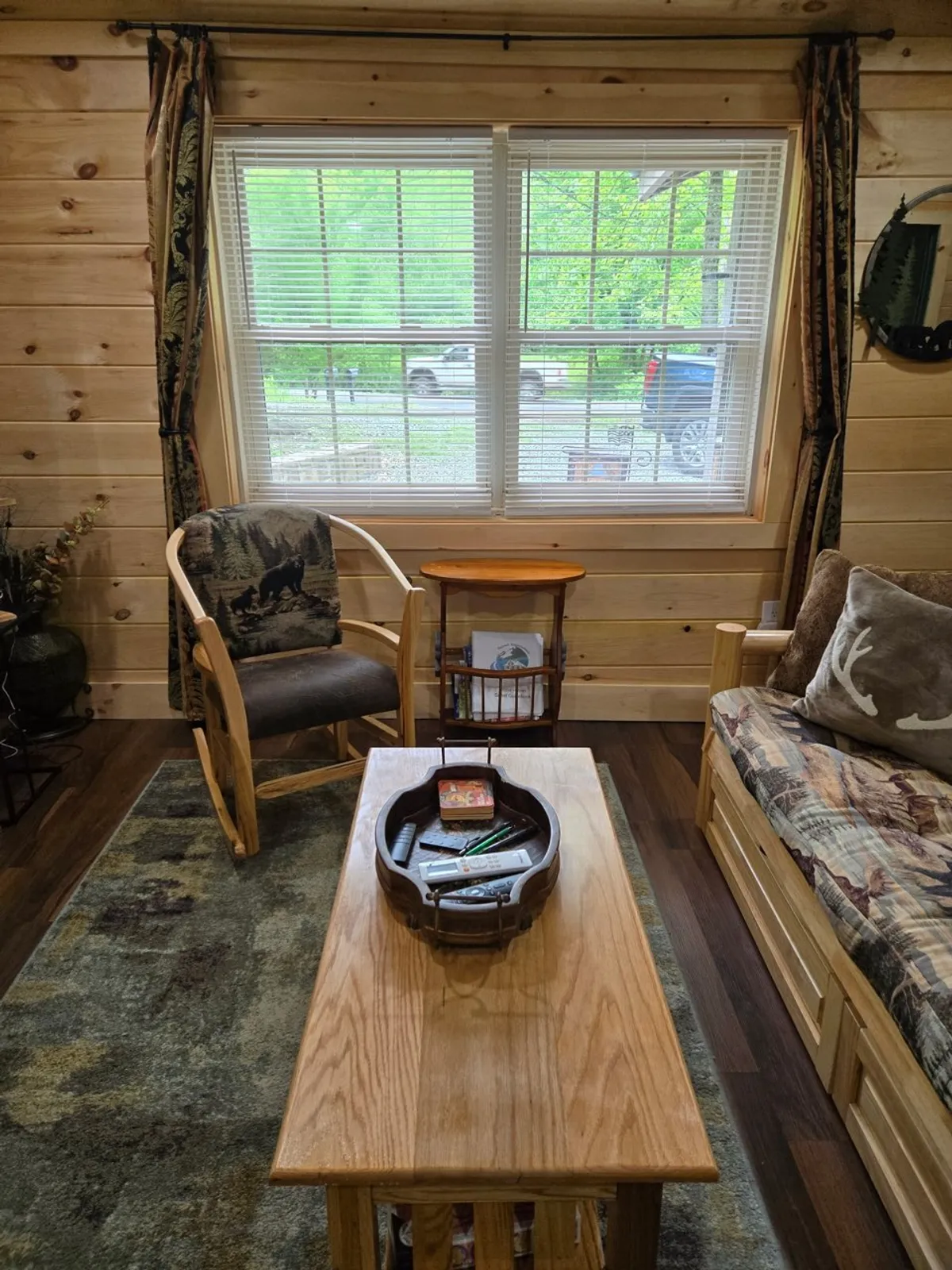 A cozy living area filled with warm, rustic furnishings, including a comfortable chair and a wooden couch with patterned cushions. Large windows showcase lush greenery just outside, inviting in natural light and offering direct visual access to the outdoors.