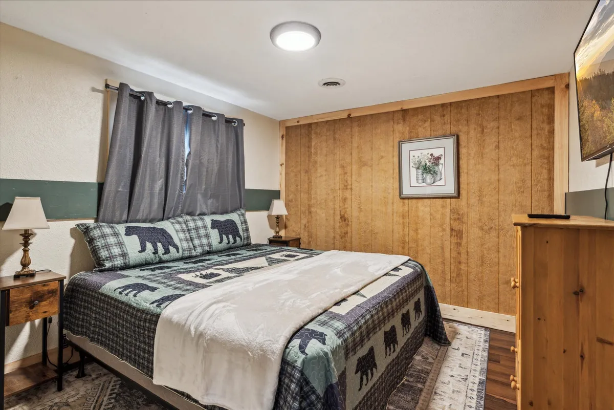 This image features a cozy master bedroom with bear-patterned bedding on a comfortable-looking bed, a wooden accent wall, a mounted television, and a nightstand with a lamp. The scene radiates warmth and invites relaxation, highlighting plush bedding and tranquil surroundings.