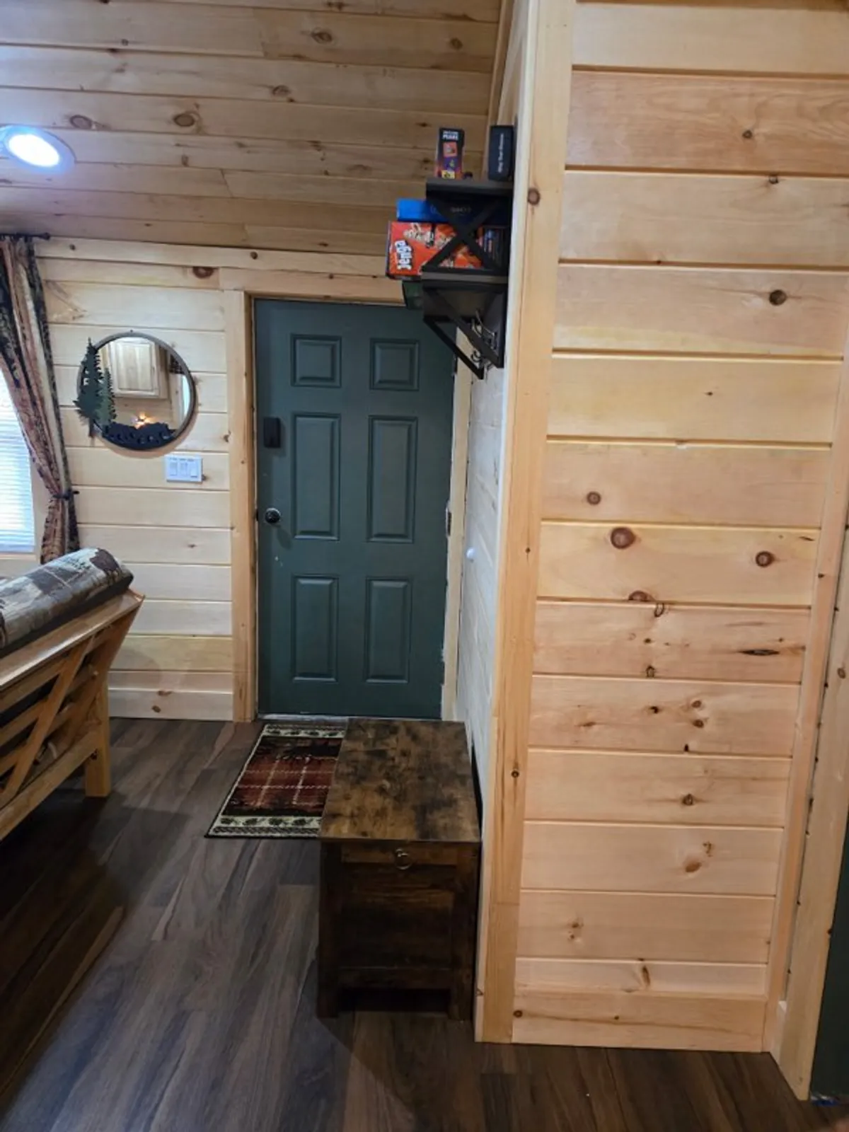 A welcoming entryway featuring a green door, wooden storage bench for outdoor gear, and a decorative tree-themed mirror. The space is functional for storing leashes or pet essentials after coming inside from an outdoor excursion.