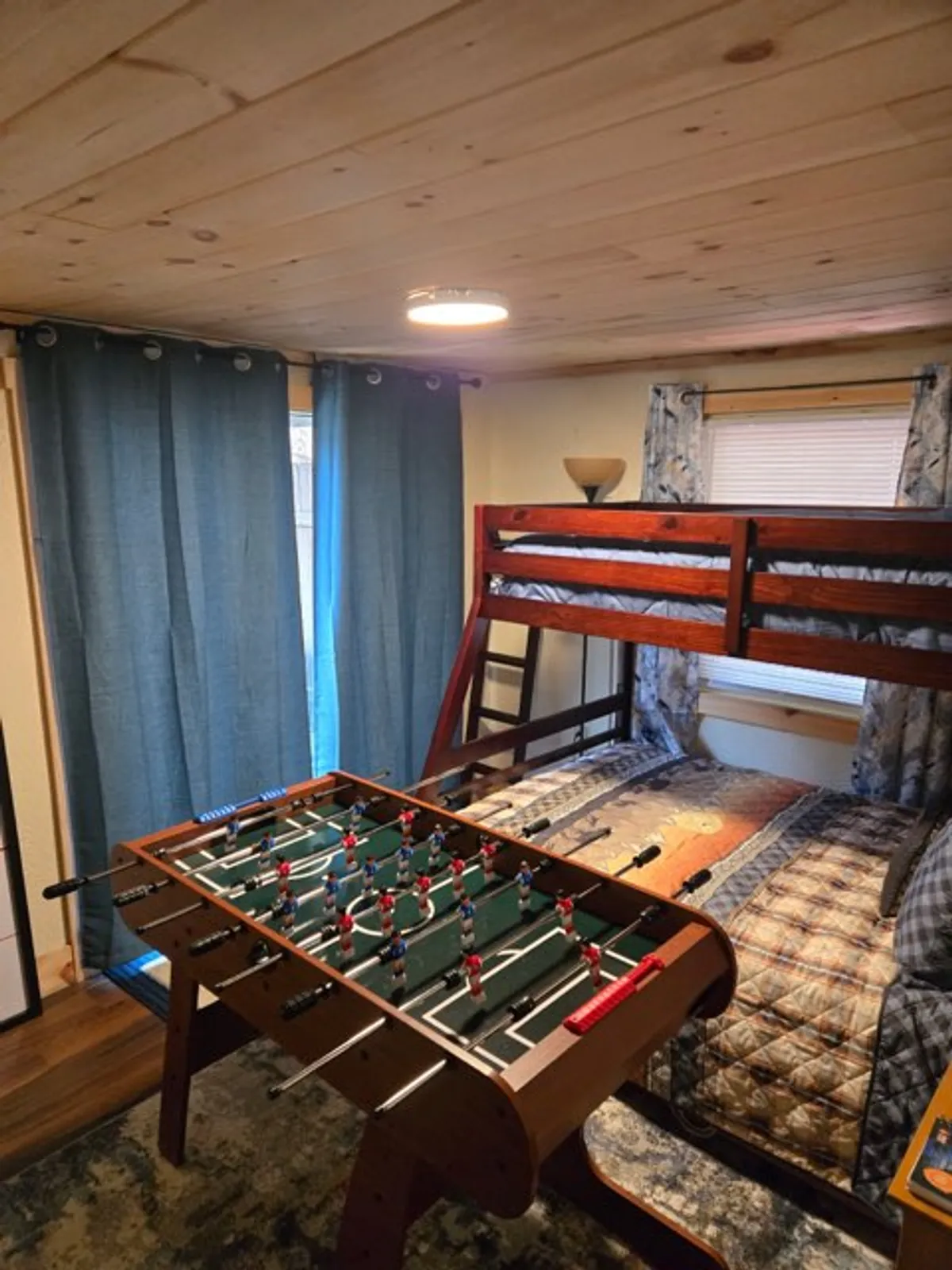 A game area featuring a foosball table, a bunk bed in the background, and blue curtains. The space is vibrant and energetic, set up for active play and socializing, ideal for family fun or friendly competition.