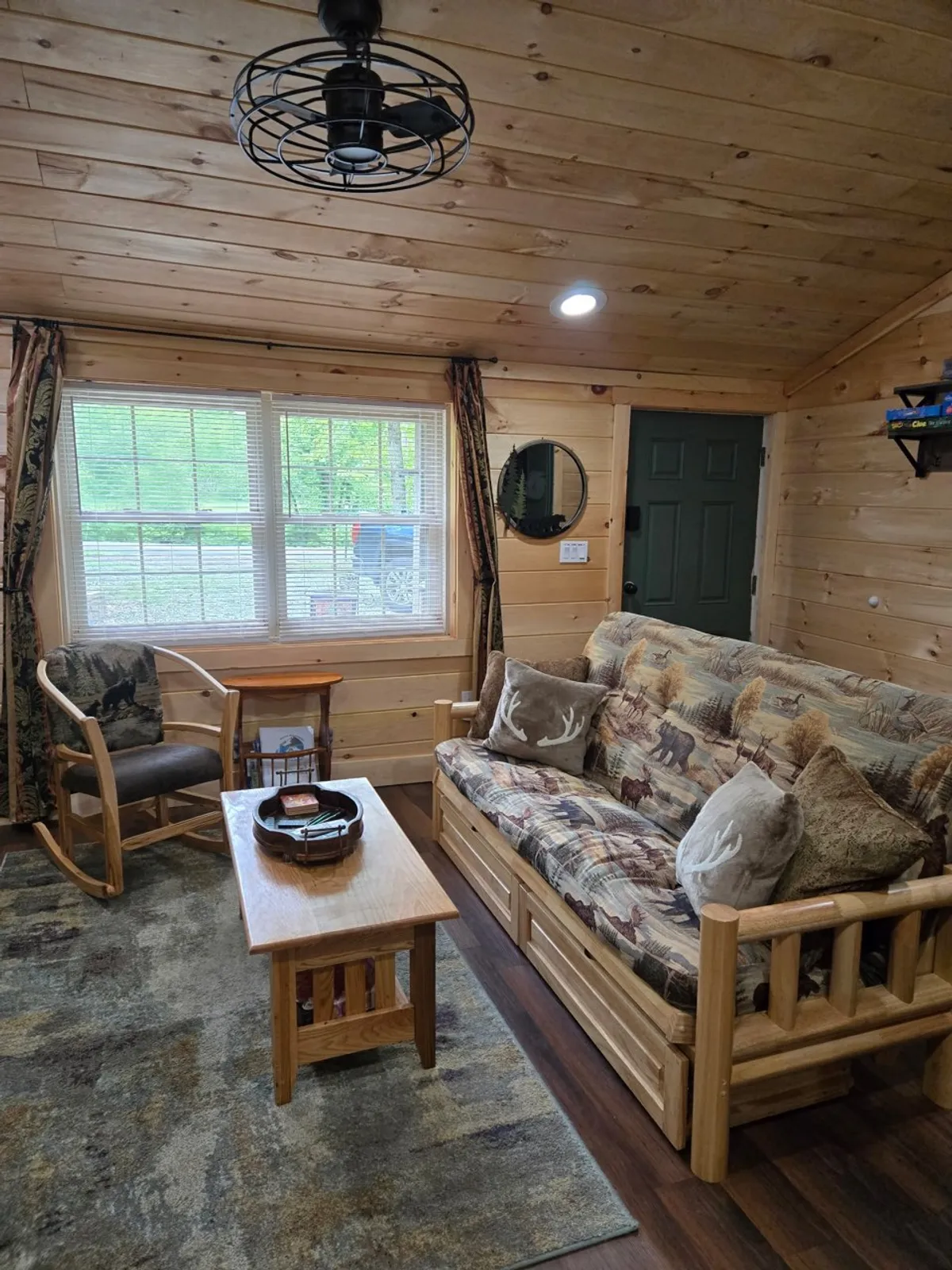 A rustic cabin living space with wooden furniture, wildlife-patterned textiles, and a large window framing a scenic outdoor view. The space feels inviting and is ideal for quiet moments, socializing, or enjoying the natural surroundings from the comfort of indoors.