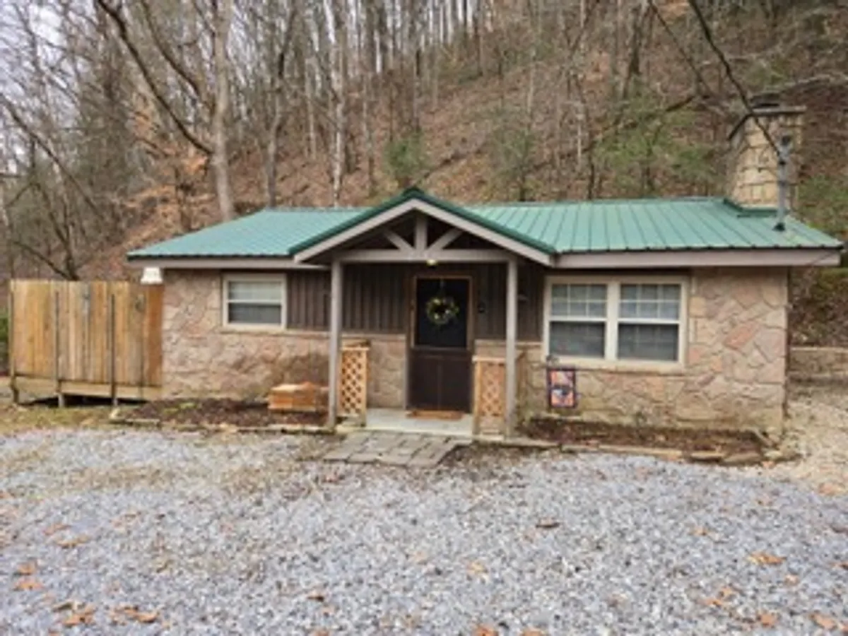 This image features the inviting front entrance of the cabin, with a welcoming wreath on the door, a charming green metal roof, and stone exterior. The porch suggests a tranquil space to relax, with hints of the surrounding natural landscape. The setting conveys the promise of peaceful mornings or romantic evenings spent together outdoors.
