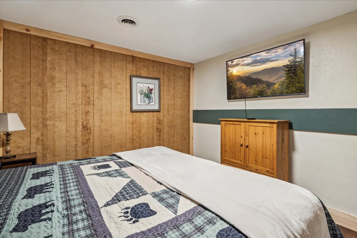 A cozy bedroom featuring a bed with a quilted cover, rustic wood paneling, framed artwork, a wall-mounted television displaying a serene landscape, and a wooden cabinet. Soft lighting and homey decor create an inviting, tranquil atmosphere ideal for unwinding.