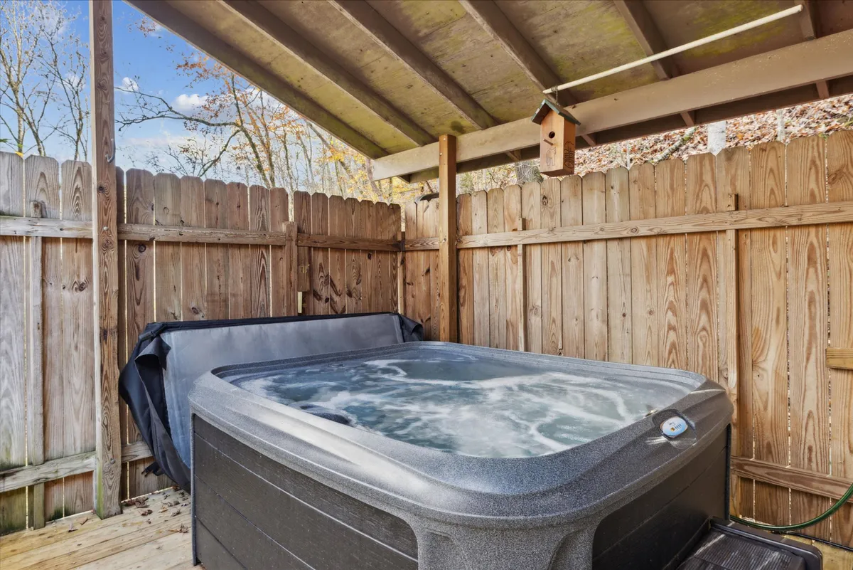 A serene view of a hot tub beneath a roof, surrounded by a wooden fence and natural greenery, with a birdhouse visible nearby. The partially covered design allows year-round enjoyment and provides a cozy, intimate setting for relaxation.